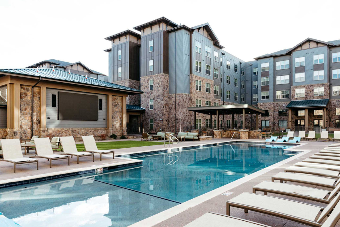 Large resort style pool with lounge chairs surrounding it and apartment building overlooking it.